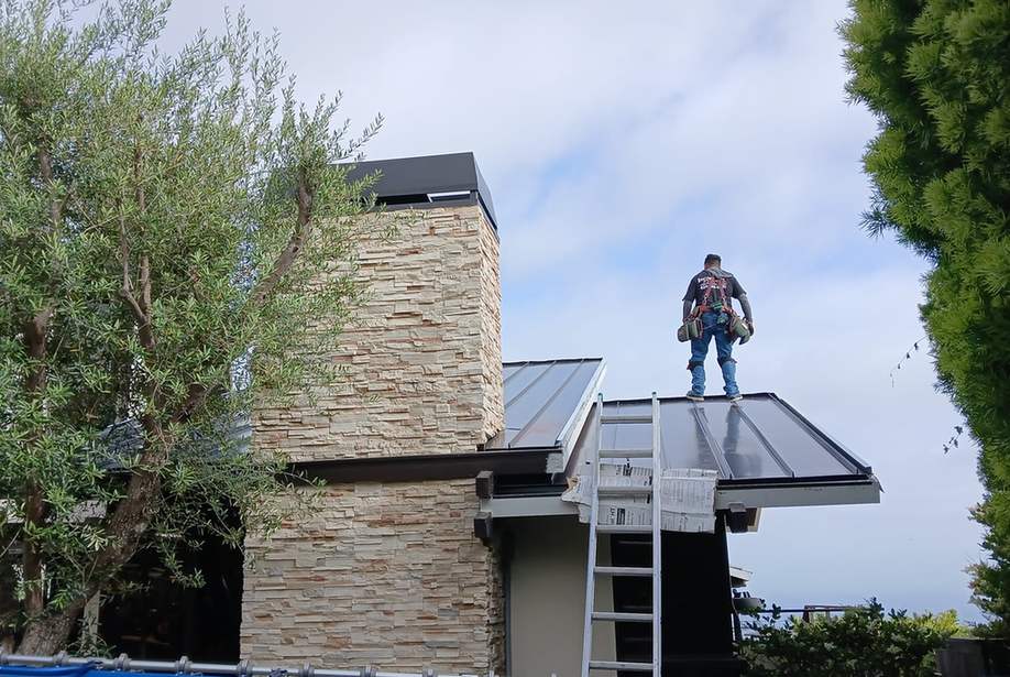 Roofer standing on a finished metal roof beside a stone chimney, featured on the Altadena roofing page.
